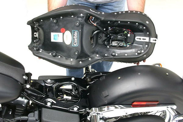 a person holding Dual Tour seat showing the rear side with Corbin Logo, Wiring harness and mounting brackets pre-installed